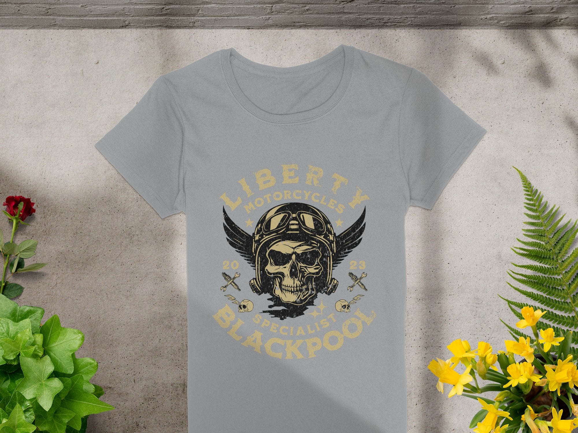A striking skull and wings design with a helmet and goggles adorns a gray t-shirt on concrete. The text Liberty Motorcycles Specialist Blackpool 2023 adds flair, complemented by nearby potted plants with yellow and red flowers—ideal for Blackpool-themed T-shirt fans. Brand: Garment Graphics.