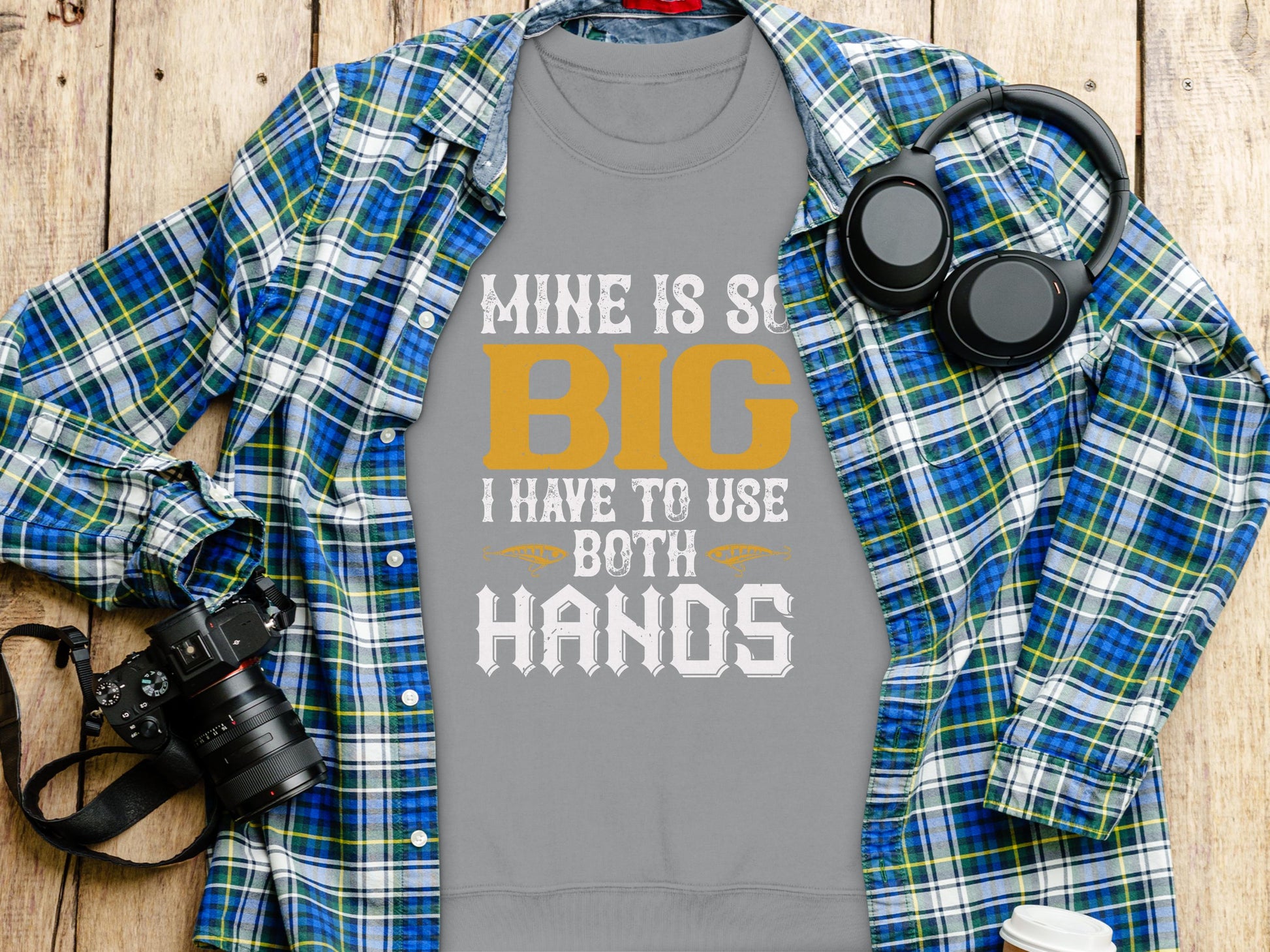 A Garment Graphics grey T-shirt with the bold text Mine is so big I have to use both hands laid flat pairs perfectly with a blue and green plaid shirt, digital camera with strap, and black headphones, all arranged on a wooden surface.