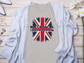 A Garment Graphics classic fit T-shirt featuring a British flag and the statement Dont blame me, I didnt vote for him lies on a wooden surface, adorned with a light gray cardigan and white flowers.