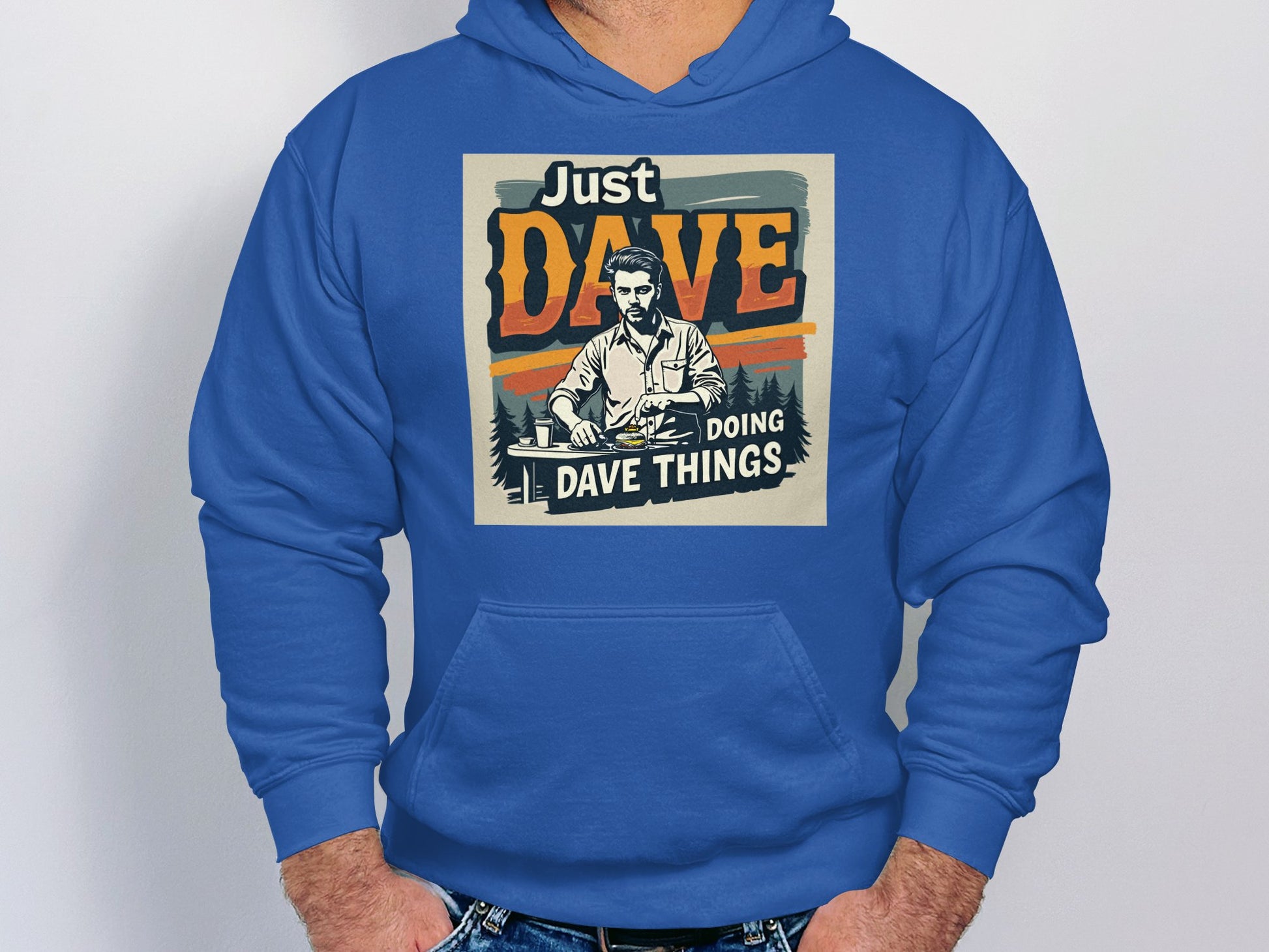 A person wears a blue Garment Graphics hoodie made from medium-heavy fabric, featuring Just Dave Doing Dave Things text and a desk graphic. Against a plain white background, their hands in jeans pockets highlight their relaxed style.