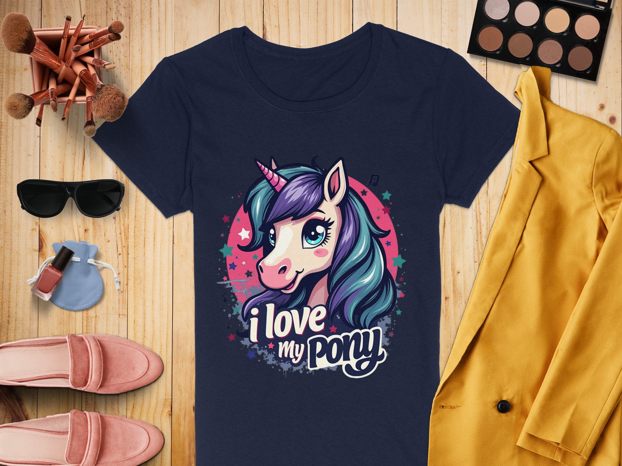 A Garment Graphics navy t-shirt with a whimsical unicorn graphic and I love my pony design rests on a wooden surface, perfect for unicorn lovers. Its surrounded by a yellow blazer, chic sunglasses, pink shoes, makeup essentials, nail polish, and a dainty purse.