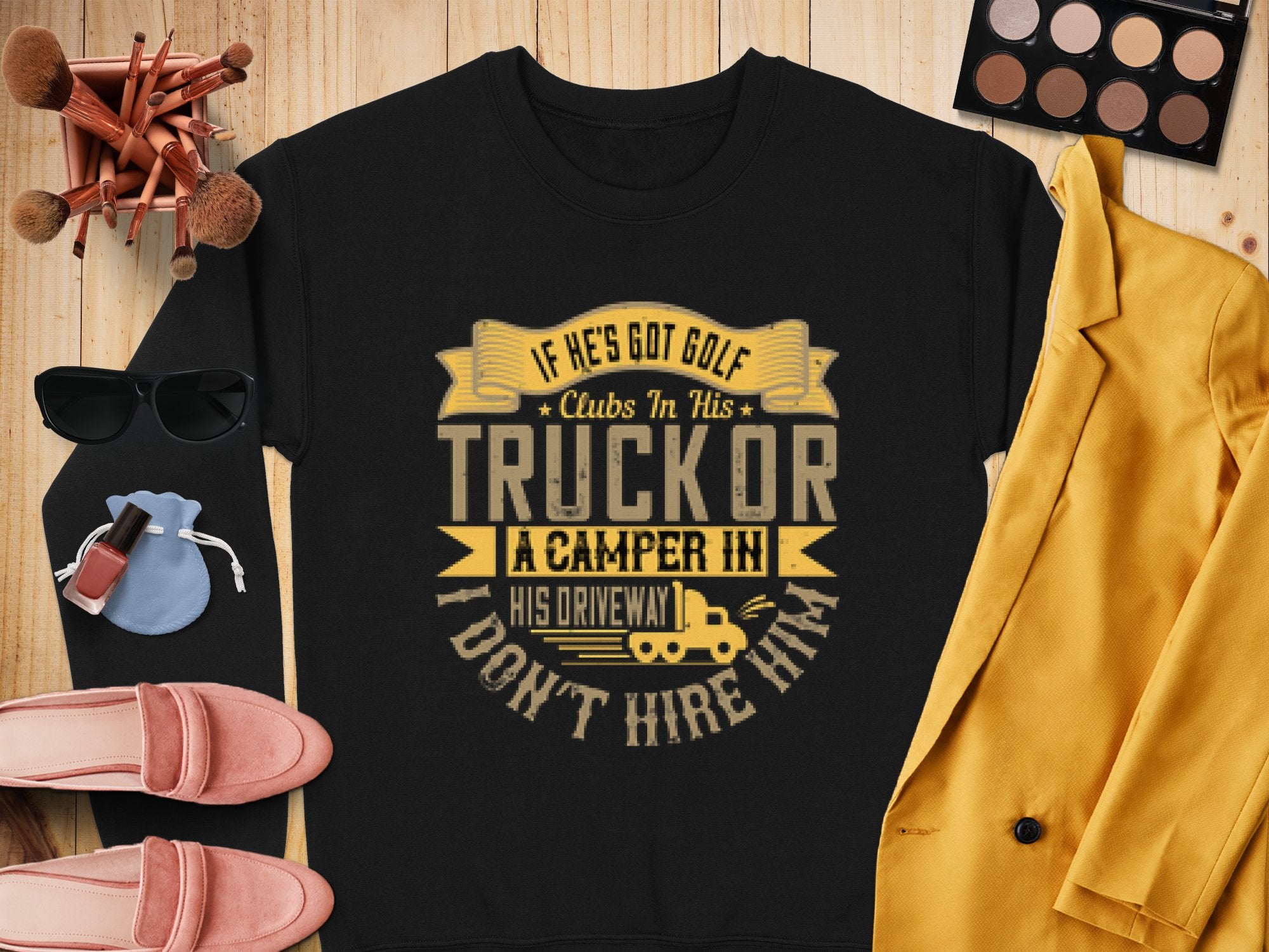 A black sweatshirt from Garment Graphics with a funny graphic saying, If hes got golf clubs in his truck or a camper in his driveway, I dont hire him, surrounded by chic items like a yellow blazer, makeup palette, brushes, sunglasses, shoes, a watch, and a small bag.