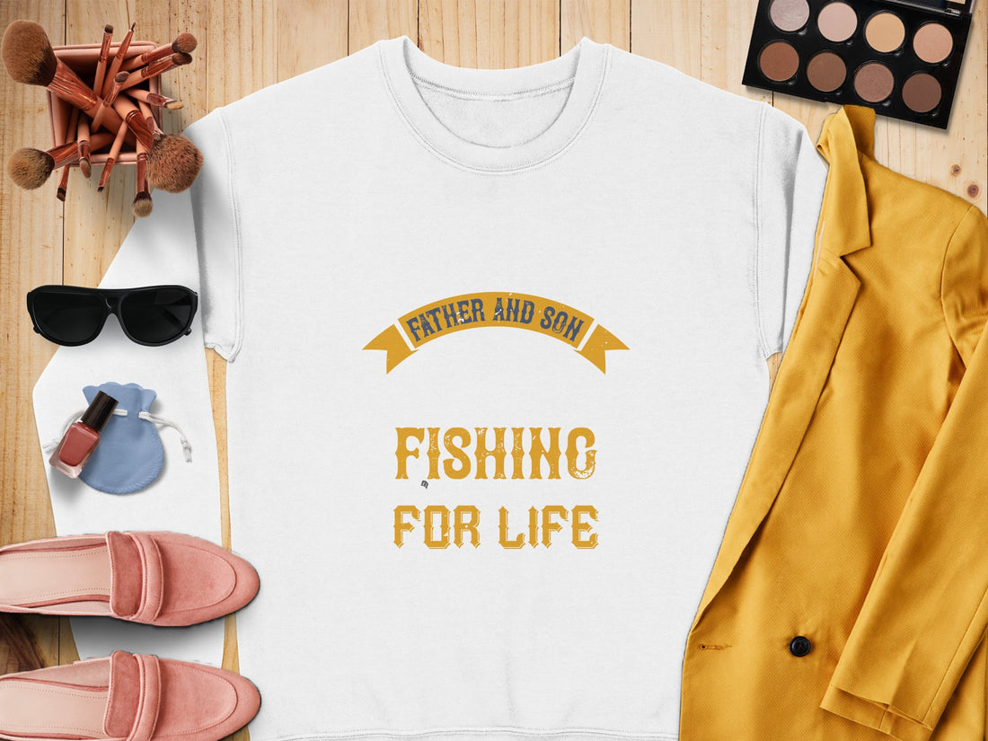 A white graphic sweatshirt by Garment Graphics, featuring Father and Son, Fishing for Life in yellow, is displayed on a wooden surface surrounded by makeup brushes, a palette, sunglasses, lipstick, a blue pouch, pink loafers, and a mustard blazer.