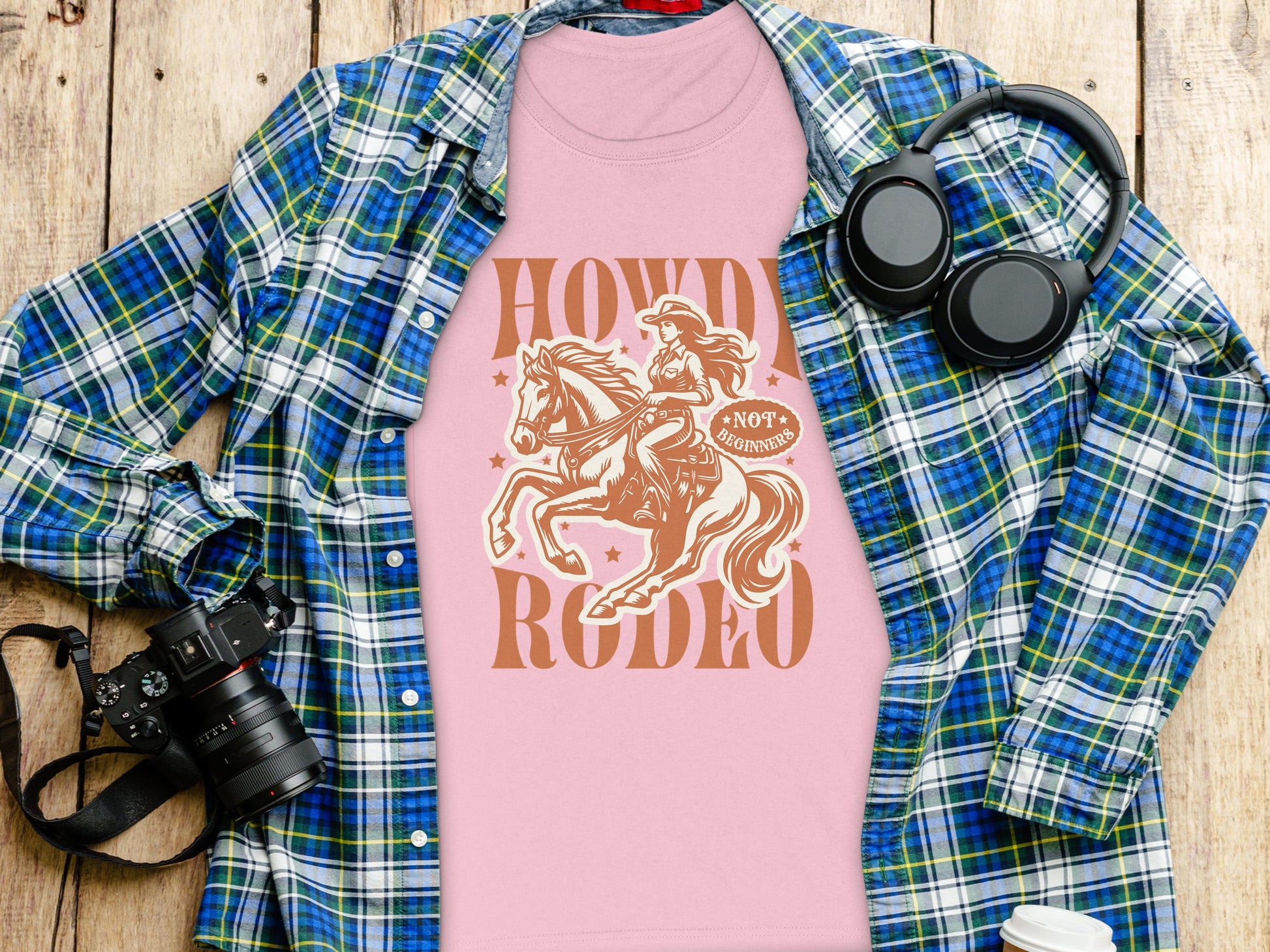 A Garment Graphics womens pink Rodeo T-Shirt with a Howdy Rodeo cowboy graphic lies on a blue and green plaid shirt. A camera, headphones, and a white cup are nearby on the wooden surface. The design shows a cowboy riding a horse with flair.