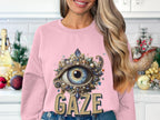 In a festive kitchen, a woman with long, wavy blonde hair wears a Garment Graphics sweatshirt featuring an ornate eye design and the word GAZE. Holiday decorations can be seen in the background.