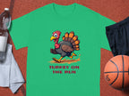 This Garment Graphics classic fit green t-shirt features a humorous turkey running and the phrase TURKEY ON THE RUN below. Perfect for sports enthusiasts, it’s displayed with a water bottle, white towel, black shorts, and a basketball on the sports surface.