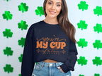 A woman stands before a clover-adorned wall wearing a cozy navy blue Garment Graphics sweatshirt, featuring Youre My Cup of Tea in bright orange. Paired with denim shorts, the outfit blends charm and comfort due to its relaxed unisex sizing.