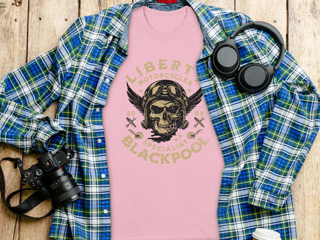 A flat lay showcases a pink Garment Graphics t-shirt with a skull and Liberty Motorcycles Blackpool 2023 Specialists print. Perfect for vintage design lovers and motorcycle enthusiasts, its surrounded by a blue-green plaid shirt, camera, and black headphones on a wooden surface.