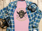 A flat lay showcases a pink Garment Graphics t-shirt with a skull and Liberty Motorcycles Blackpool 2023 Specialists print. Perfect for vintage design lovers and motorcycle enthusiasts, its surrounded by a blue-green plaid shirt, camera, and black headphones on a wooden surface.
