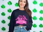 In front of a wall with green shamrocks, a woman sports a black sweatshirt from Garment Graphics showcasing a pink graphic of three figures and the phrase Always Party Beach Girl. She pairs it seamlessly with denim shorts for a relaxed, festive vibe.