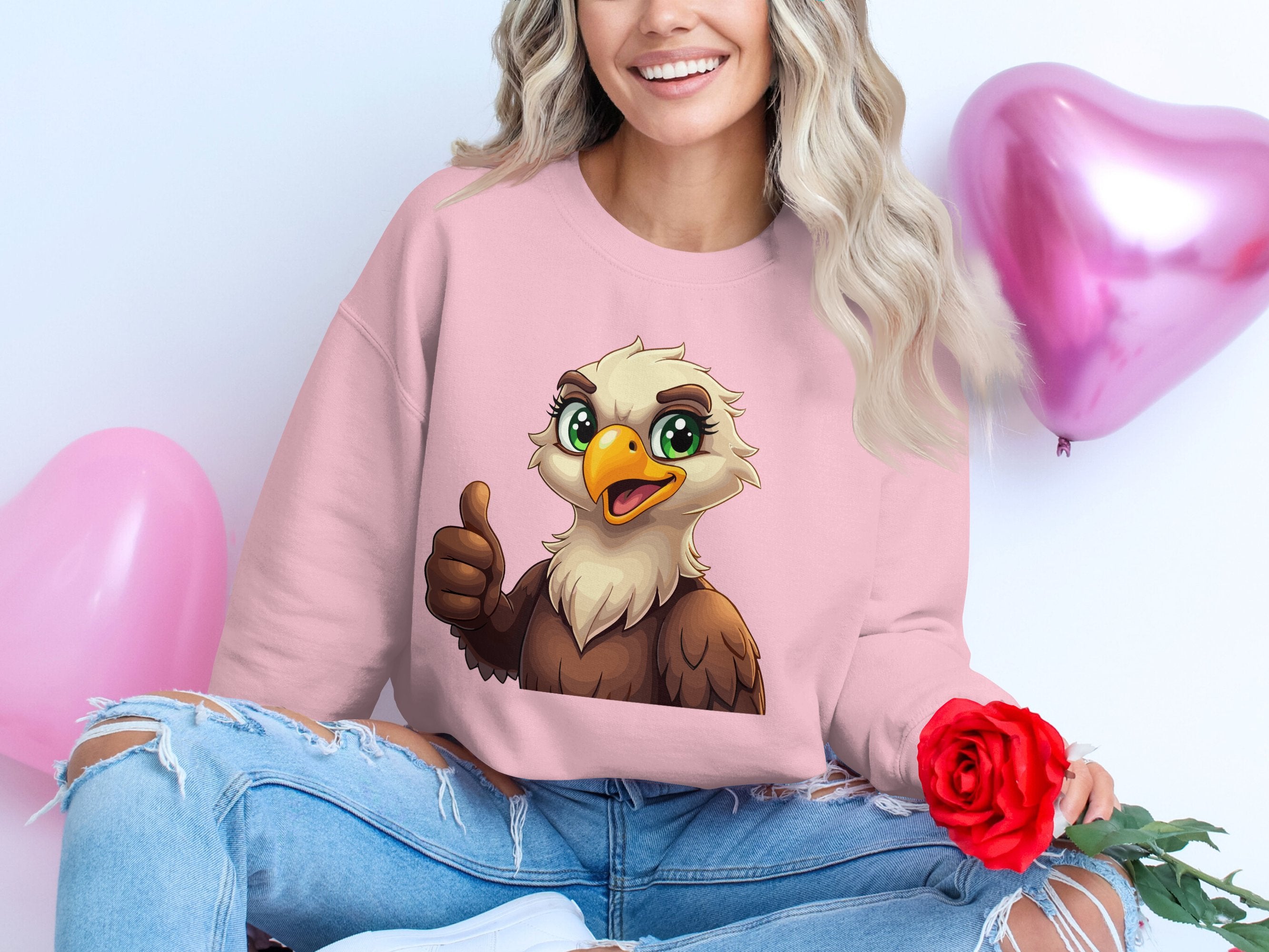 A person smiles while sitting cross-legged in a cheerful, cartoon eagle sweatshirt by Garment Graphics, featuring unisex sizing. They hold a red rose, and two pink heart-shaped balloons float behind them.