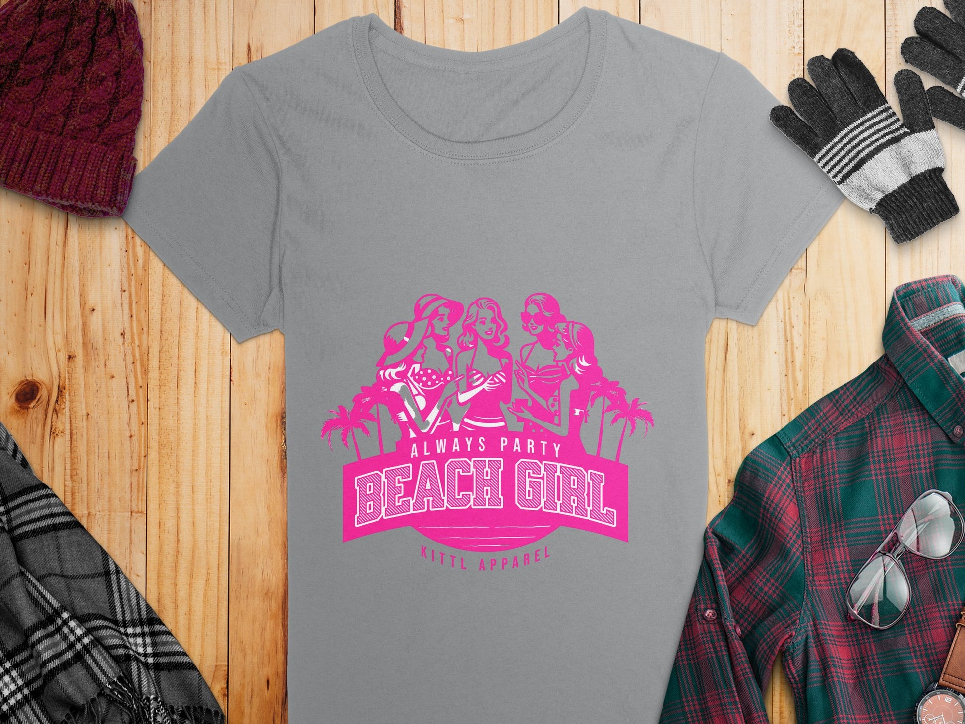 A gray Womens Semi-Fitted Tee from Garment Graphics features a pink print with three cartoon figures and drinks, framed by palm trees, and text saying Always Party Beach Girl. Displayed on wood with a hat, gloves, and plaid clothing.