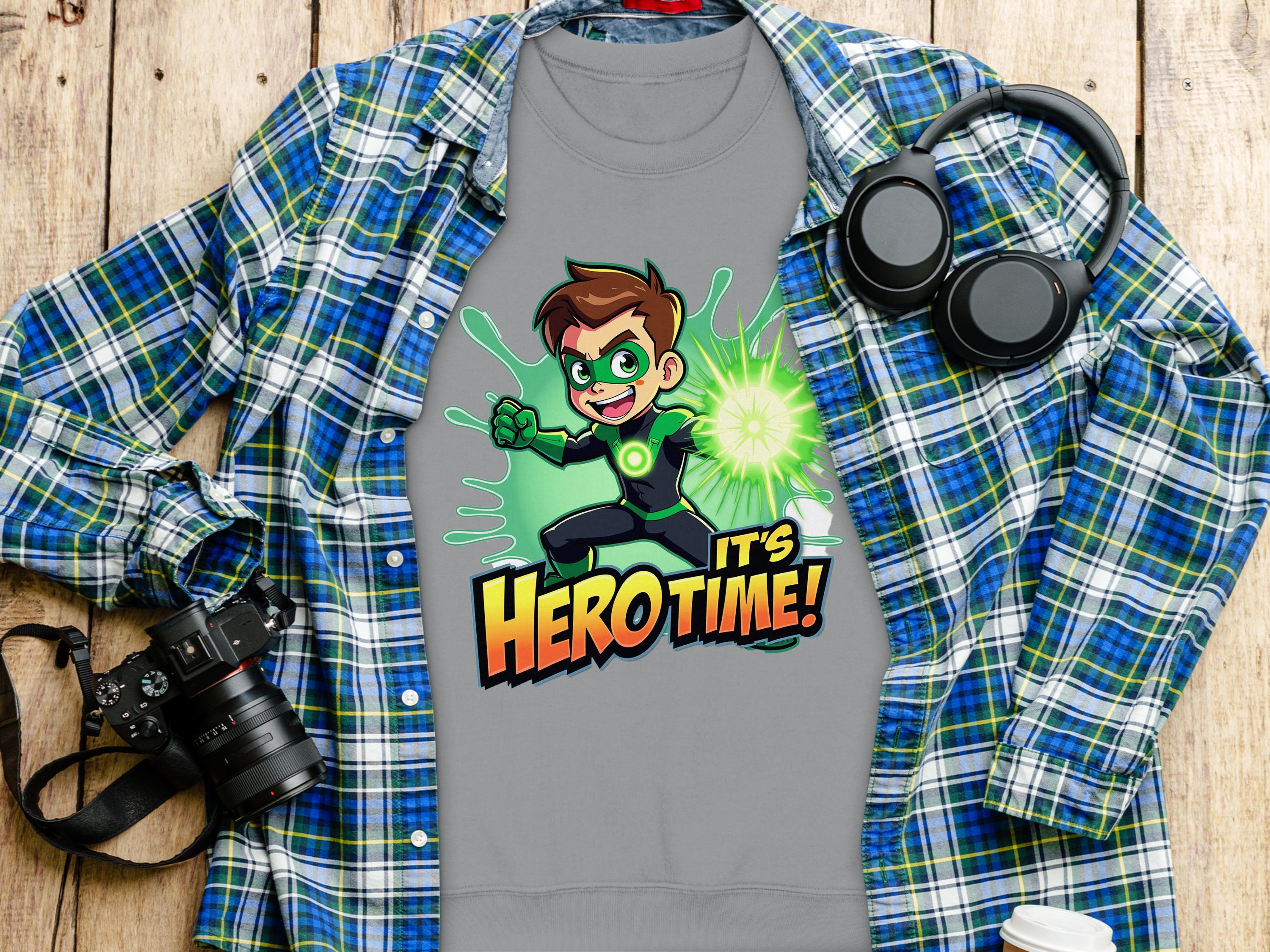 The Garment Graphics gray T-shirt featuring an Its Herotime character in a green outfit is perfect for adventurous youth, artistically displayed on a blue and green plaid shirt beside a black camera, headphones, and white cup on a wooden surface.