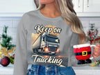 A person with long hair wears a gray unisex sweatshirt by Garment Graphics, featuring a truck and the text Keep on Trucking. They hold a Santa outfit-themed mug amidst festive Christmas decorations.