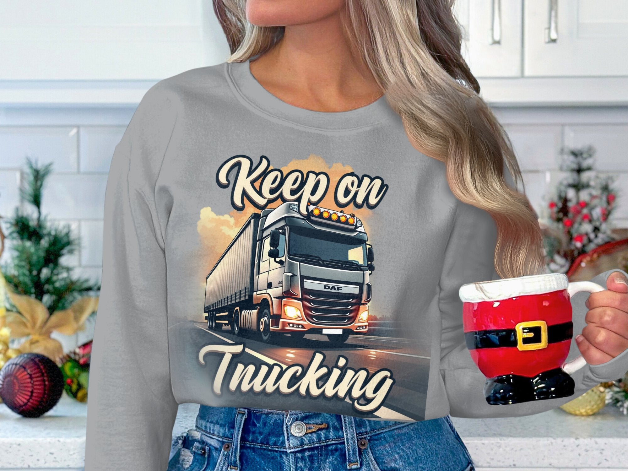 A person with long hair wears a gray unisex sweatshirt by Garment Graphics, featuring a truck and the text Keep on Trucking. They hold a Santa outfit-themed mug amidst festive Christmas decorations.