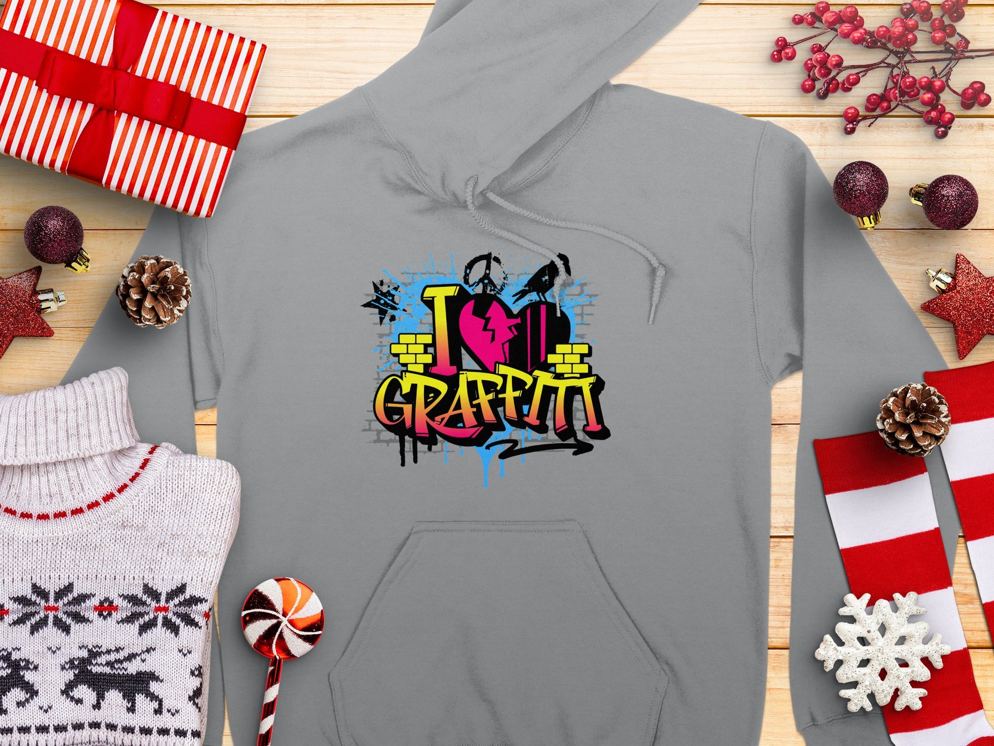 The Garment Graphics gray hoodie features a colorful I Love Graffiti design with paint cans and a brick wall, perfect for casual wear. Its surrounded by holiday items like wrapped gifts, pinecones, red berries, a red star ornament, candy cane, snowflake, and a patterned sweater.