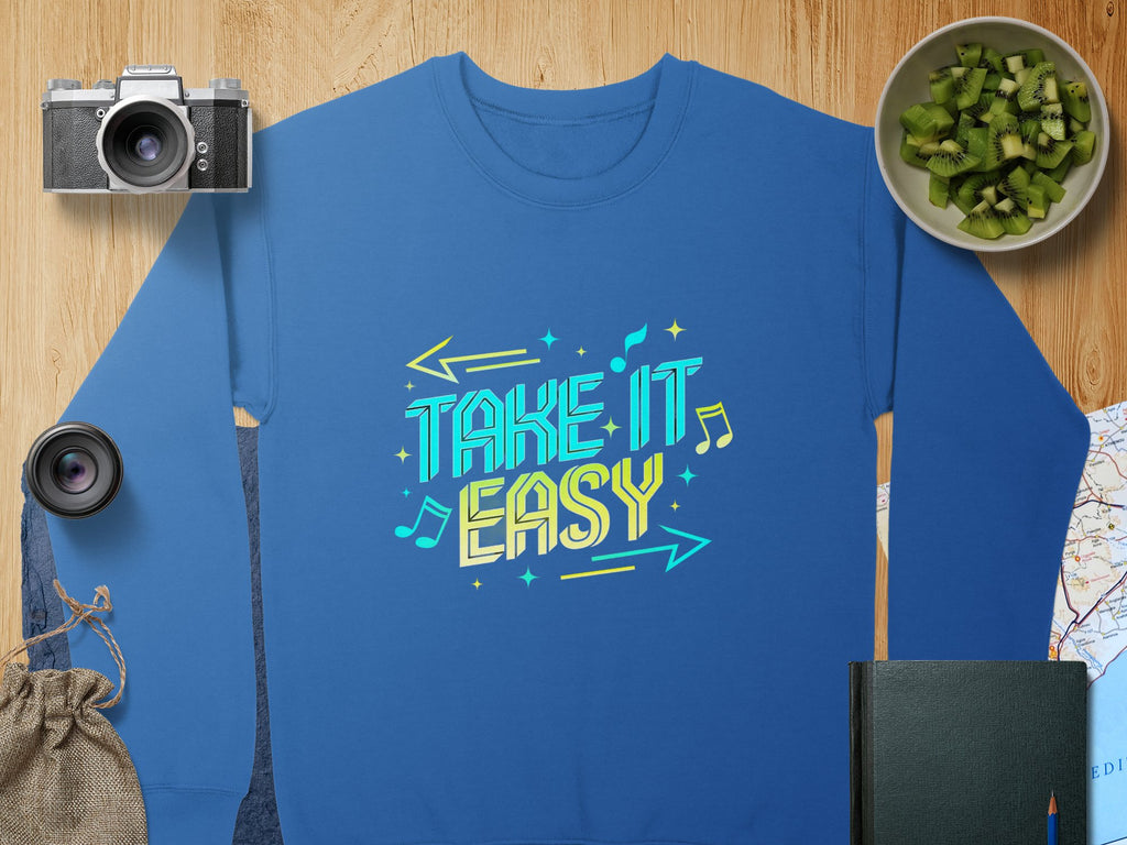 The Garment Graphics Unisex Sweatshirt in blue showcases a vibrant Take It Easy graphic in yellow and white. Styled on a wooden surface with a camera, lens, sliced kiwis, notebook, map, and burlap pouch for the ultimate casual ensemble.