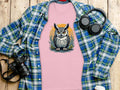 A pink Garment Graphics owl graphic T-shirt adds artistic flair beneath a blue and green plaid shirt on a wooden surface, adjacent to black headphones, a camera, and a white bottle.