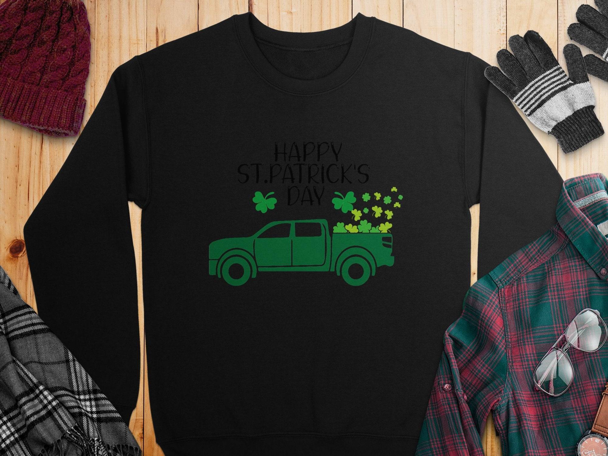 Garment Graphics festive sweatshirt showcases a green truck packed with shamrocks and Happy St. Patricks Day text, surrounded by a red beanie, gloves, black-and-white scarf, and green plaid shirt on wood.