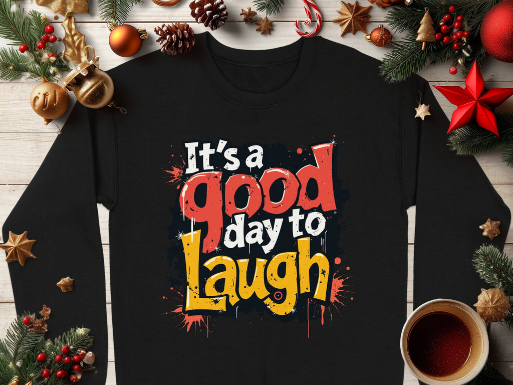 A medium-heavy black shirt from Garment Graphics with Its a good day to laugh in colorful letters lies on wood surrounded by pine cones, orange slices, and a red star. Perfectly unisex, it pairs beautifully with a cozy cup of coffee or tea for anyone.