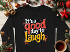 A medium-heavy black shirt from Garment Graphics with Its a good day to laugh in colorful letters lies on wood surrounded by pine cones, orange slices, and a red star. Perfectly unisex, it pairs beautifully with a cozy cup of coffee or tea for anyone.