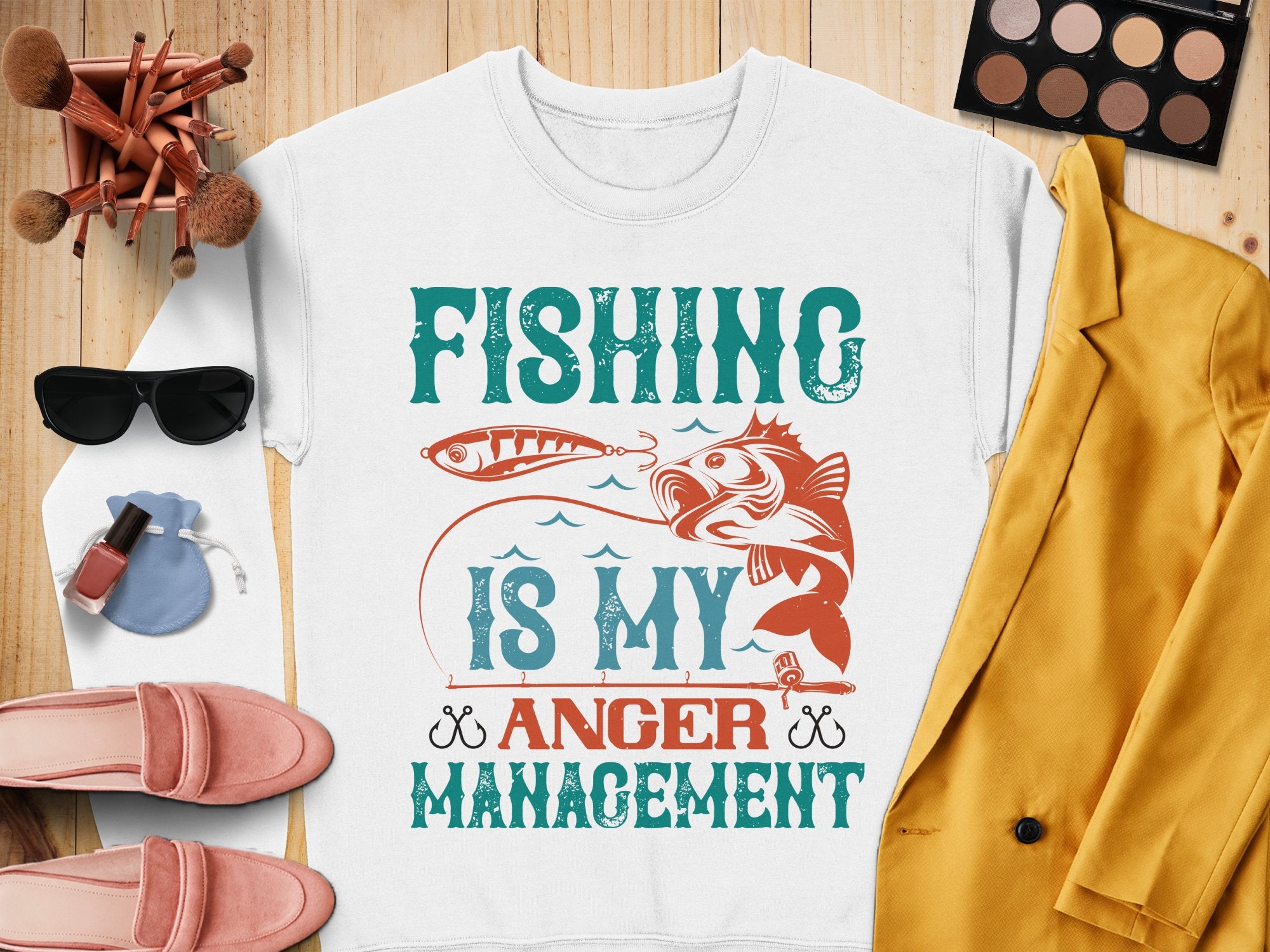 Garment Graphics presents a white T-shirt featuring Fishing Is My Anger Management with a fish and hook design, styled alongside a yellow jacket, pink shoes, sunglasses, makeup brushes, nail polish, and an eyeshadow palette on a wooden surface. Perfect for fishing enthusiasts.