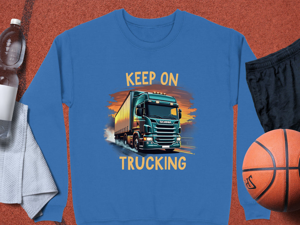 A Garment Graphics blue graphic sweatshirt with a bold truck design and KEEP ON TRUCKING text is displayed on a white towel beside a water bottle, black shorts, and an orange basketball.