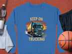 A Garment Graphics blue graphic sweatshirt with a bold truck design and KEEP ON TRUCKING text is displayed on a white towel beside a water bottle, black shorts, and an orange basketball.