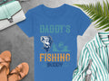 Garment Graphics blue Daddys Fishing Buddy t-shirt, with a boat and fish design, is paired with tan sandals, a striped shirt, watch, light blue purse, and green ferns on concrete—perfect for an avid anglers casual day out.