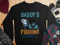 Garment Graphics black fishing sweatshirt showcases Daddys Fishing Buddy in playful blue, orange, and teal. It features a person fishing from a boat with a big catch on the line, surrounded by winter wear on wood—a must-have for fishing fans tackling chilly outings.