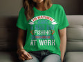 A person is sitting on a sofa wearing Garment Graphics green fishing T-shirt that reads, Id rather have a bad day fishing than a good day at work, ideal for fishing enthusiasts.