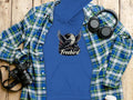 Garment Graphics blue Freebird Graphic Pullover Hoodie showcases an eagle in flight under a green and blue plaid shirt. Paired on a wooden surface with headphones, a camera, and a white lidded cup, its the ideal ensemble to highlight your unique style.