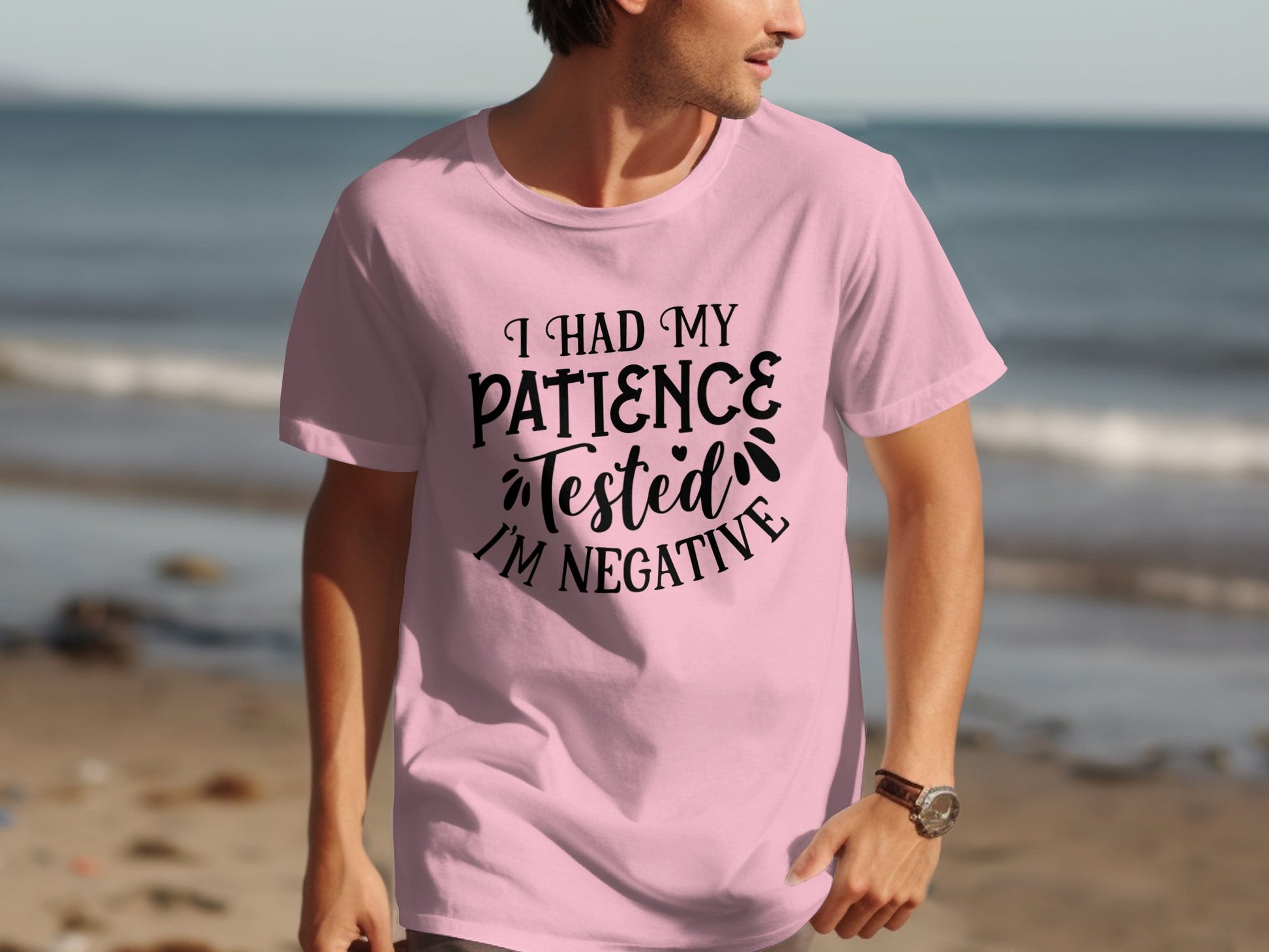 Wearing a light pink Garment Graphics T-shirt reading I Had My Patience Tested, Im Negative, a person strolls along the beach. The scene, set against sand, water, and a partly cloudy sky, perfectly matches their sarcastic vibe.