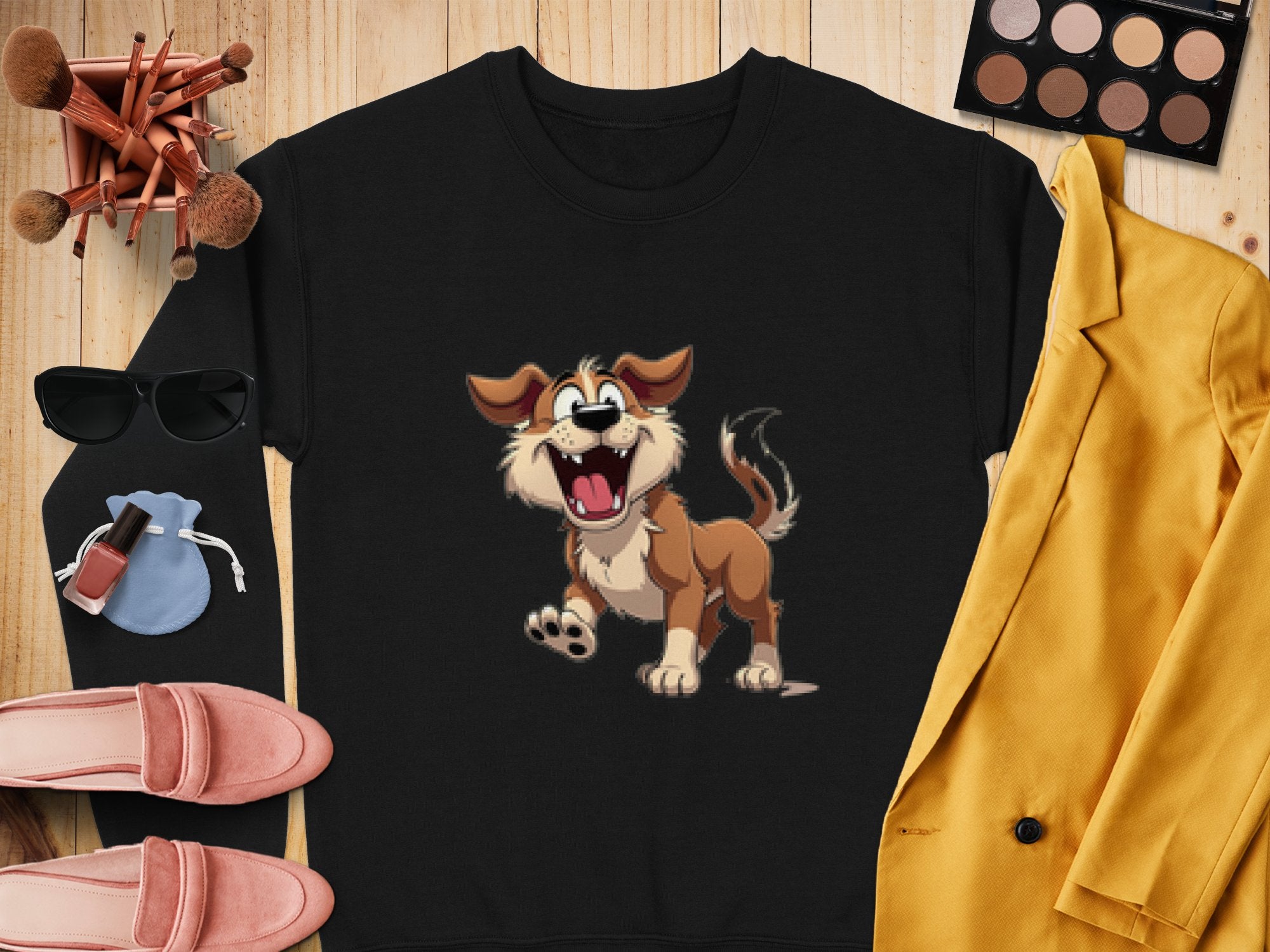 Garment Graphics black sweatshirt with a happy dog illustration sits beside yellow jacket, makeup brushes, eyeshadow palette, pink loafers, and blue pouch on wood—ideal for dog lovers.