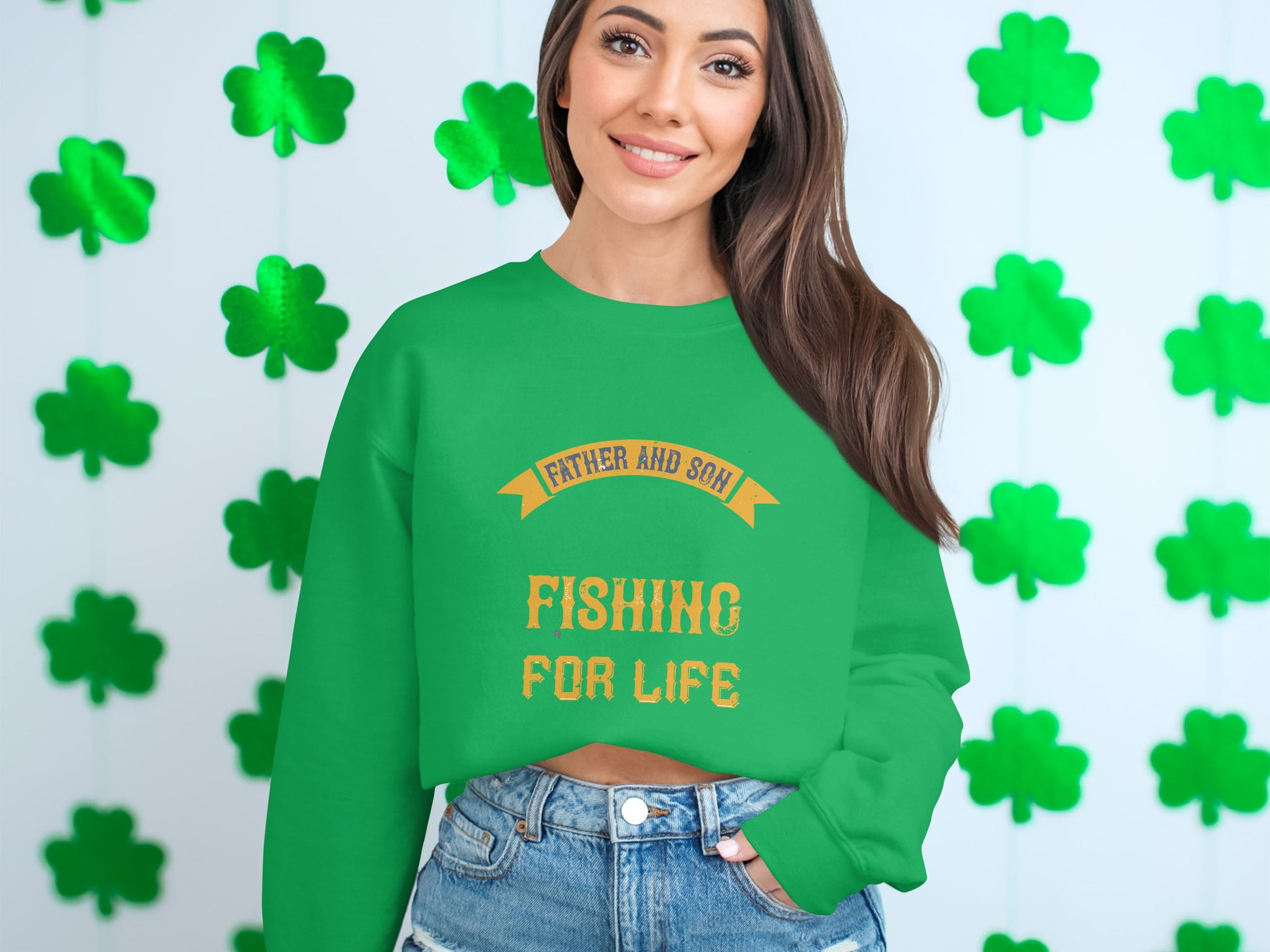 Against a backdrop of green shamrocks, a person wears a vibrant Garment Graphics sweatshirt reading Father and Son Fishing for Life in bright yellow. Paired with denim shorts, they smile warmly at the camera, capturing the joy of family bonds and shared passions.