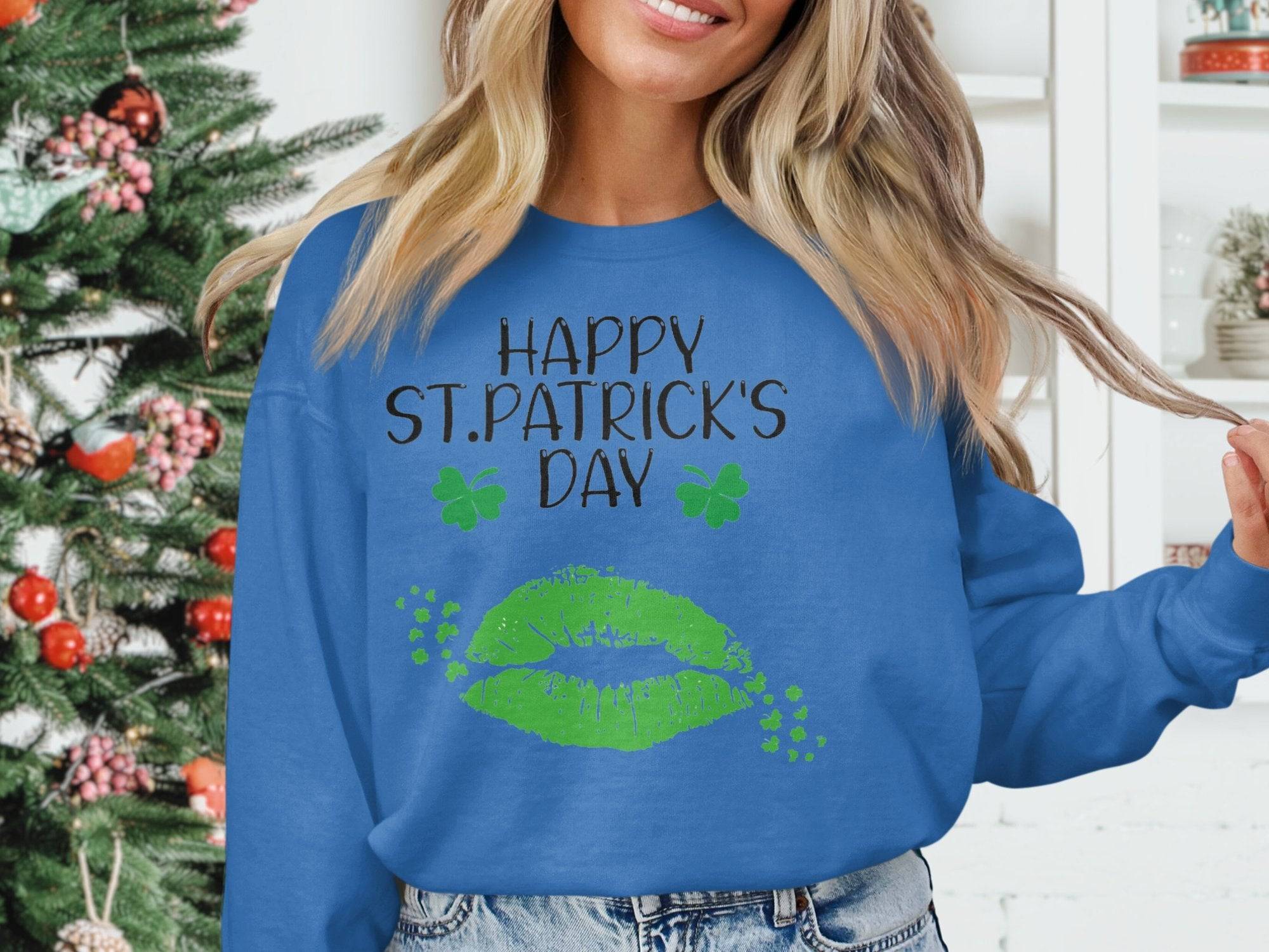 A person in a blue Garment Graphics sweatshirt featuring Happy St. Patricks Day and clover kiss designs stands near a decorated Christmas tree, embracing the festive spirit.