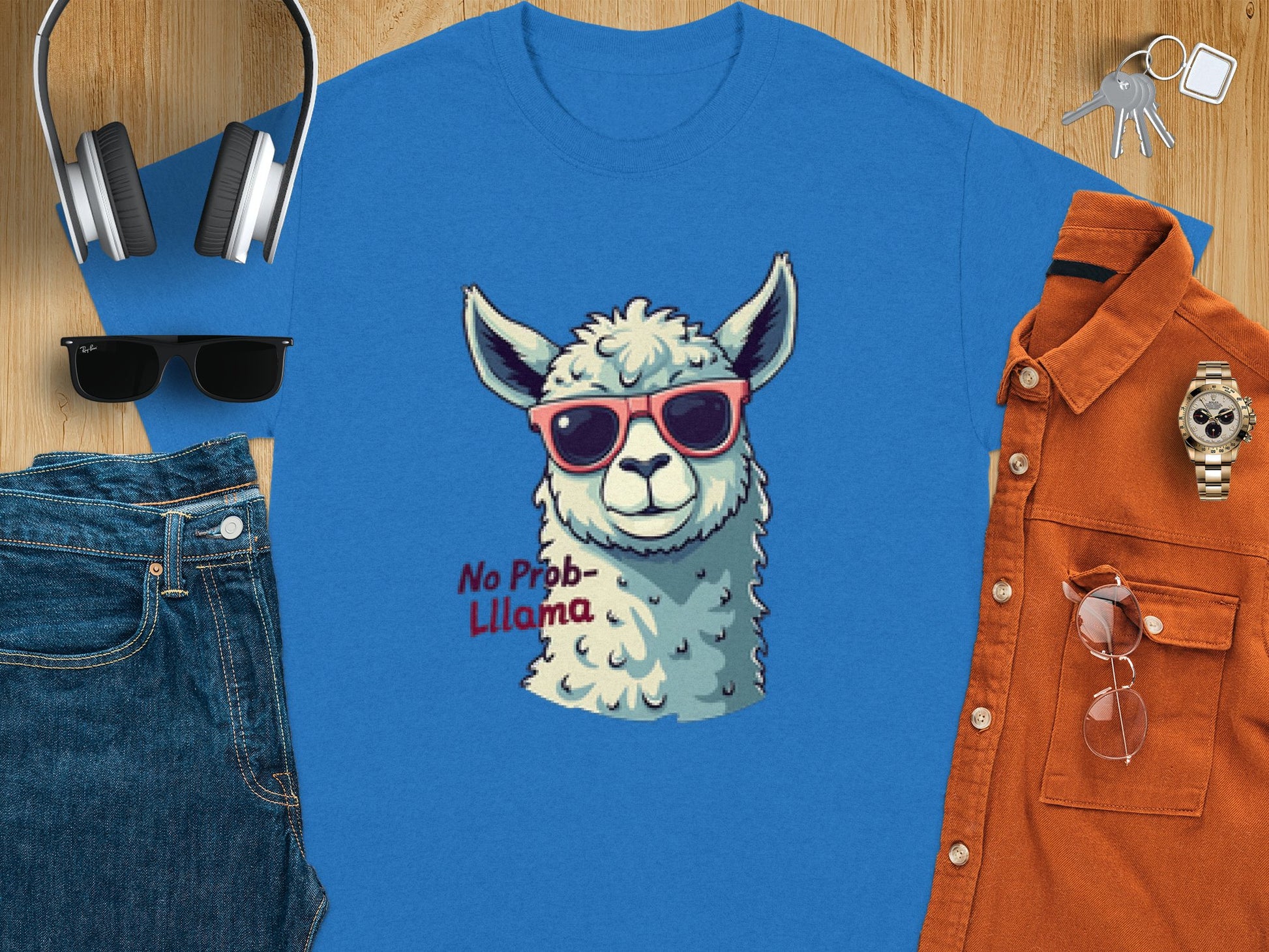 Garment Graphics Blue Llama T-shirt features a stylish llama in sunglasses with No Prob-Llama text. Nearby are headphones, keys, additional sunglasses, a watch, an orange shirt, and blue jeans on wood. Ideal for family sizing adventures!.