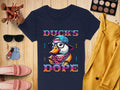 A navy blue T-shirt by Garment Graphics features a Cool Brave Wild Swag Duck with a cap and glasses stating Ducks Dope, surrounded by makeup brushes, an eyeshadow palette, sunglasses, headphones, nail polish, pink shoes, and a yellow jacket on a wooden surface.