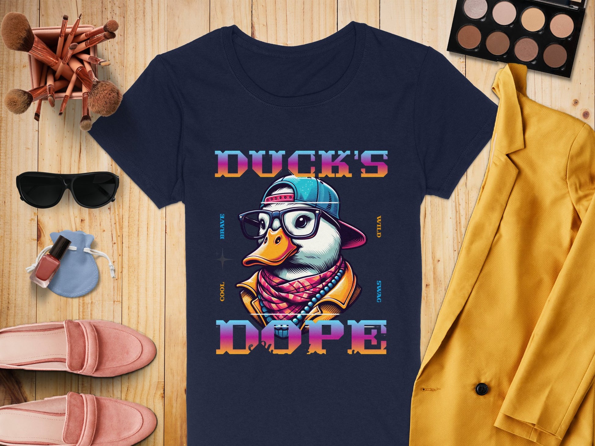 A navy blue T-shirt by Garment Graphics features a Cool Brave Wild Swag Duck with a cap and glasses stating Ducks Dope, surrounded by makeup brushes, an eyeshadow palette, sunglasses, headphones, nail polish, pink shoes, and a yellow jacket on a wooden surface.