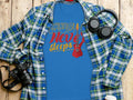 A blue graphic T-shirt featuring Music Never Sleeps with a red guitar design lies on a wooden surface beside a blue and green plaid shirt, headphones, 100% cotton comfort, a camera, and a coffee cup lid. Brand: Garment Graphics.