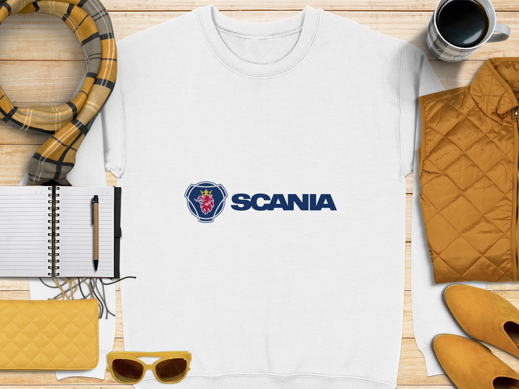 A white unisex Garment Graphics Scania t-shirt is neatly placed on a wooden surface with a pen and notebook. Its accompanied by a yellow quilted jacket, coffee cup, plaid scarf, yellow sunglasses, and matching clutch—a perfect setup for casual outings.