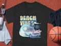 A black Garment Graphics T-shirt features a classic car, surfboards, palm trees, and BEACH VIBES text. Its displayed on a reddish surface with a basketball, black shorts, water bottle, and gray towel.