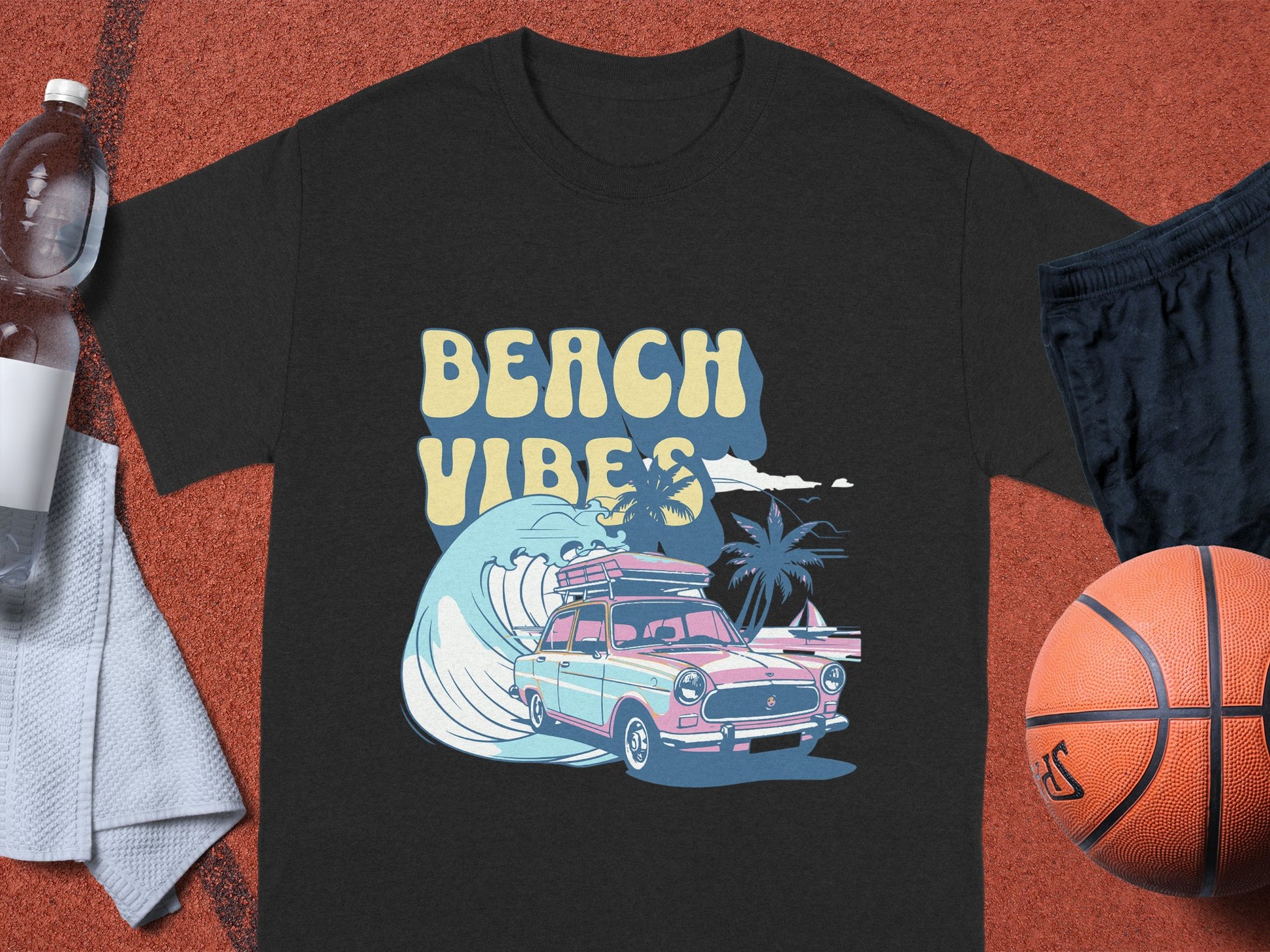 A black Garment Graphics T-shirt features a classic car, surfboards, palm trees, and BEACH VIBES text. Its displayed on a reddish surface with a basketball, black shorts, water bottle, and gray towel.