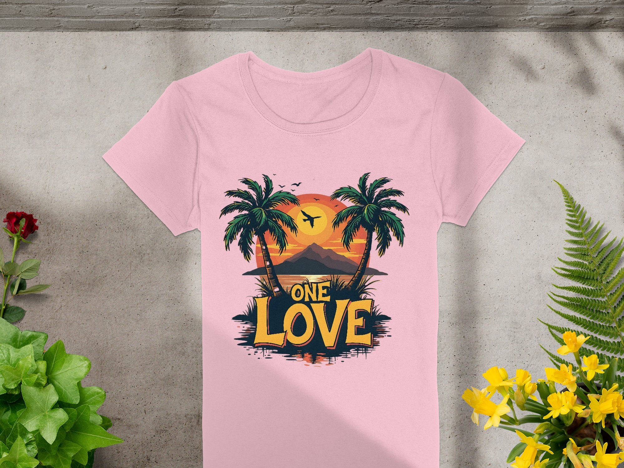 The Garment Graphics pink T-shirt is ideal for beach outings, with a design of palm trees, a tropical sunset, and a mountain. Featuring the bold slogan One Love, its displayed on concrete amidst green and yellow potted plants.