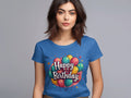 A woman stands against a gray background wearing a vibrant blue 100% cotton t-shirt by Garment Graphics, featuring a colorful birthday balloon design and the words Happy Birthday, ideal for any birthday celebration.