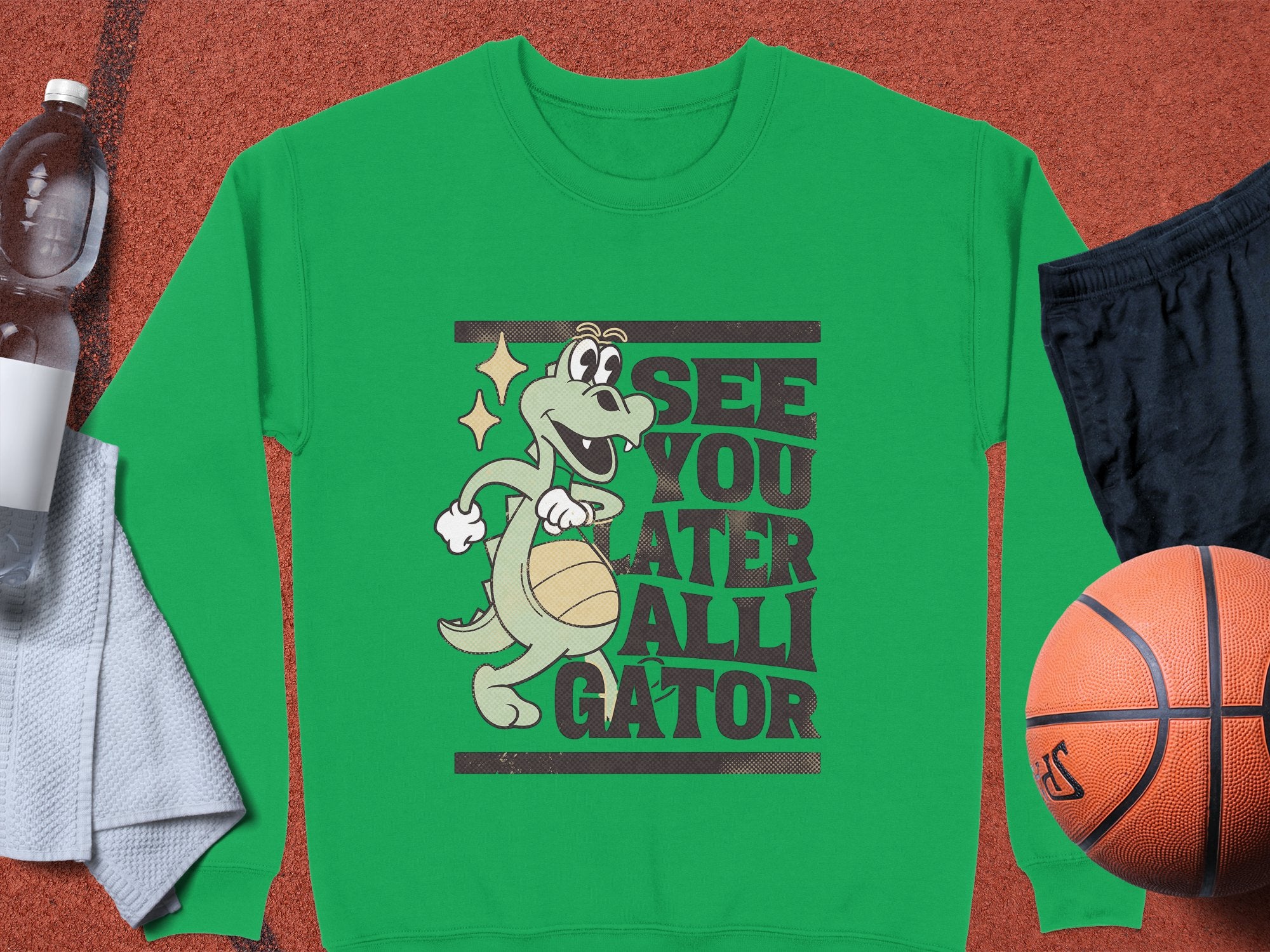 Versatile unisex sizing enhances this Garment Graphics green sweatshirt with a cartoon alligator holding a basketball. See You Later Alligator is written boldly on it, and its set against an orange background with a basketball, black shorts, water bottle, and white towel.