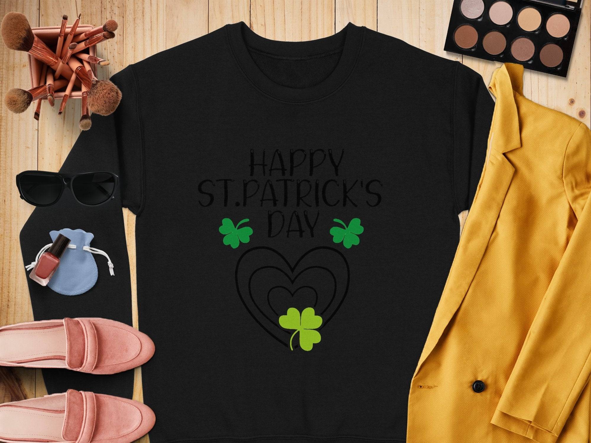 A Garment Graphics festive sweatshirt featuring Happy St. Patricks Day and a clover design sits on wood alongside a yellow jacket, pink loafers, blue pouch, makeup brushes, sunglasses, a compact, and nail polish.