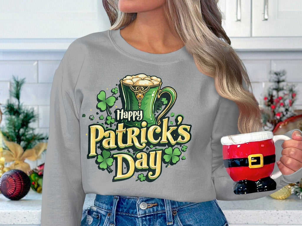 In a festive kitchen, someone wears a vibrant Garment Graphics Happy Patricks Day sweater with shamrocks and a green mug design. They hold a Santa-themed mug, mixing holiday graphics amid joyful decor.