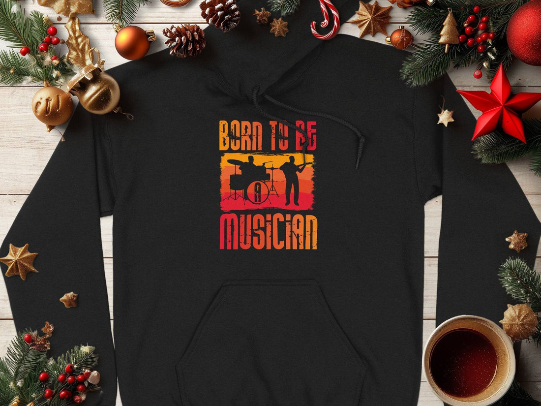 A black Garment Graphics hoodie on a wooden surface, ideal for music lovers, is adorned with an orange and red band member graphic and Born to Be a Musician text. Holiday decorations surround it, while a coffee cup enhances its cozy vibe. Unisex fashion at its best.