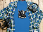 A blue, 100% cotton Freebird eagle T-shirt by Garment Graphics with a bold graphic is beneath a blue and green plaid shirt. Nearby are headphones, a camera, and a white cup, all set on a wooden surface.